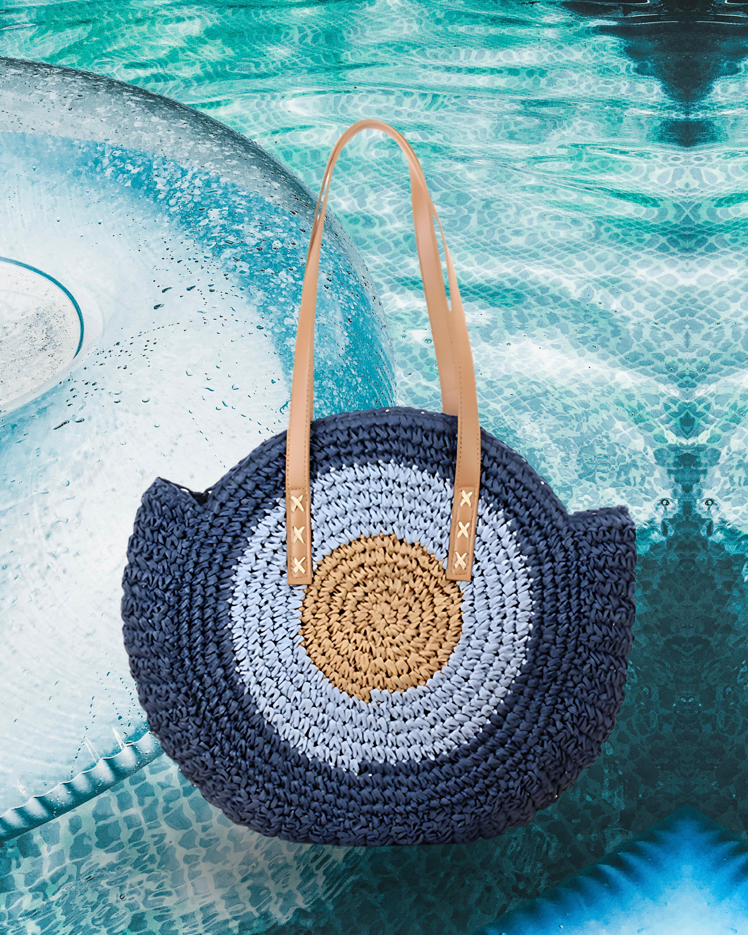 Round woven raffia tote bag with concentric tan, light blue, and navy circle design, structured shell-like silhouette, and long brown leather shoulder straps