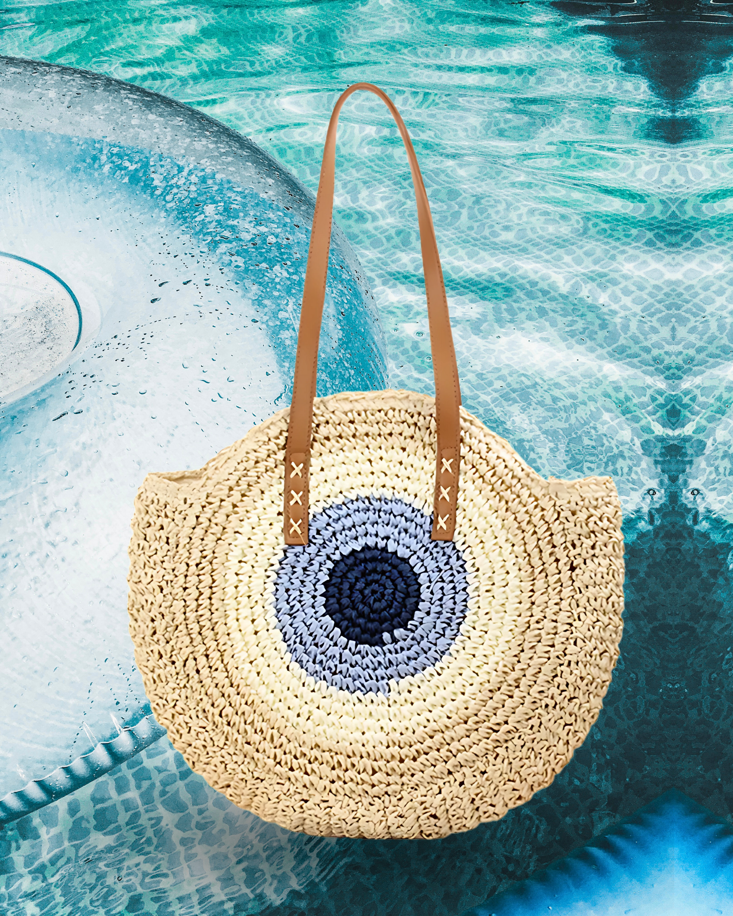 Round woven raffia tote bag with an evil eye design in navy, light blue, beige including long brown leather shoulder straps.